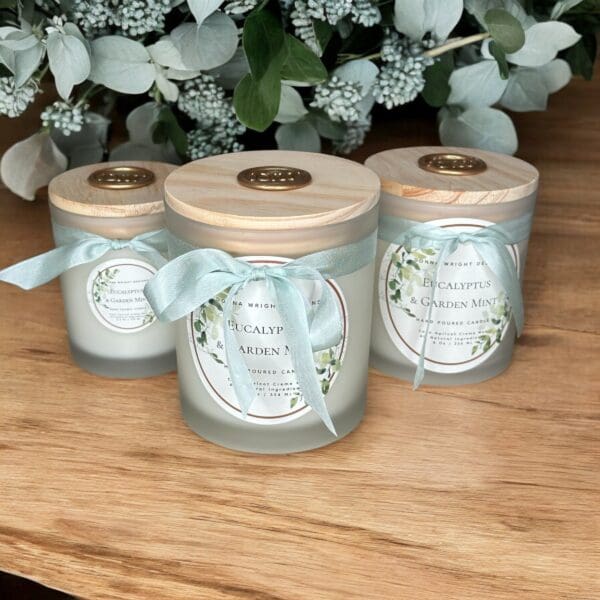 Three eucalyptus and garden mint scented candles with frosted glass jars and wooden lids are displayed on a wooden surface. Each candle is adorned with a light blue ribbon and a detailed label. Eucalyptus leaves and white flowers are in the background.