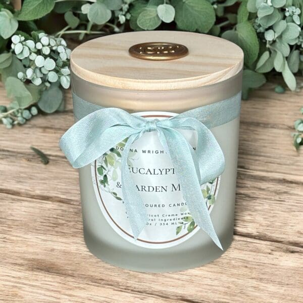 A frosted glass candle with a wooden lid is adorned with a light blue ribbon. The label reads "Eucalyptus & Garden Mint" and is decorated with illustrated leaves. The candle is placed on a wooden surface with greenery in the background.