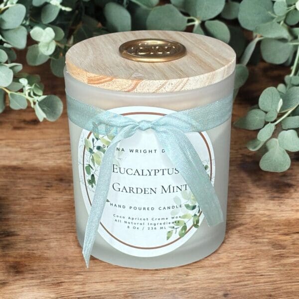 A frosted glass candle labeled "Eucalyptus Garden Mint" is placed on a wooden surface. The candle has a wooden lid with a gold emblem and is tied with a light blue ribbon. Green eucalyptus leaves surround the candle in the background.