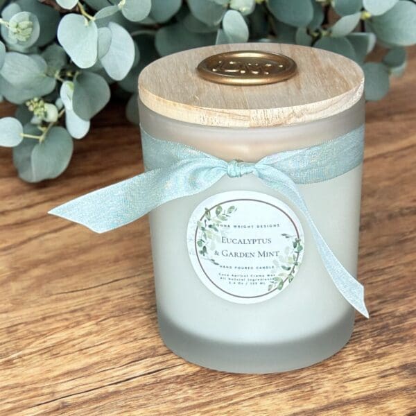 A frosted glass candle with a wooden lid sits on a wooden surface. The candle has a light blue ribbon tied around its neck and a label that reads, "Eucalyptus & Garden Mint." There are sprigs of eucalyptus in the background.