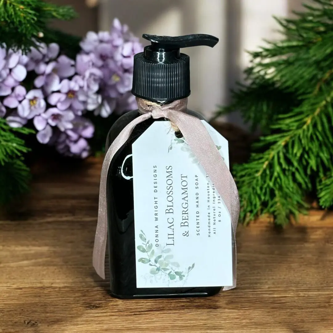A decorative bottle of scented hand soap is adorned with a brown ribbon and white label reading "Lilac Blossoms & Bergamot." The bottle is positioned on a wooden surface with purple flowers and green foliage in the background.