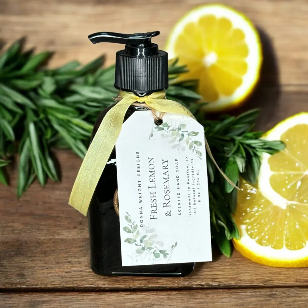 Donna Wright Designs A dark pump bottle labeled "Fresh Lemon & Rosemary Scented Hand Soap" with a yellow ribbon and gift tag displays on a wooden surface. Fresh rosemary sprigs and two lemon slices are arranged beside the bottle, enhancing the natural theme.