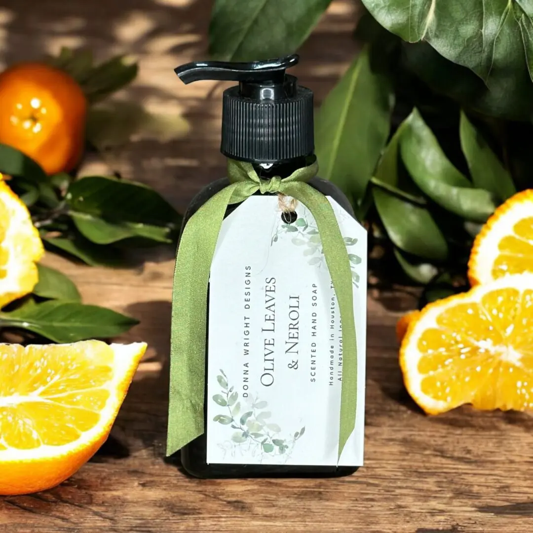 A bottle of scented hand soap with a label reading "Olive Leaves & Neroli" is adorned with a green ribbon and set against a background of greenery and orange slices. The product by Donna Wright Designs is stylishly presented with a natural, refreshing aesthetic.