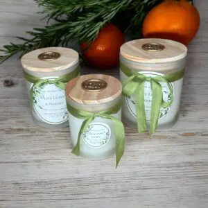 Donna Wright Designs This image shows three scented candles in frosted glass jars with wooden lids and green ribbons. The labels read "Olive Leaves & Neroli." They are placed on a wooden surface with a sprig of greenery and oranges in the background.