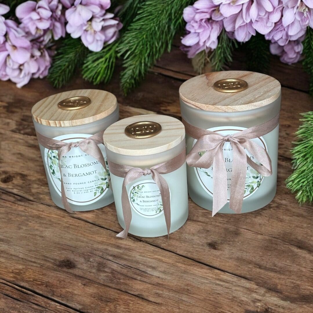 Three scented candles in frosted glass jars with wooden lids and beige ribbons are displayed on a wooden surface. The labels read "Lilac Blossom" in a floral design. They are surrounded by purple flowers and green foliage, creating a rustic and elegant ambiance.