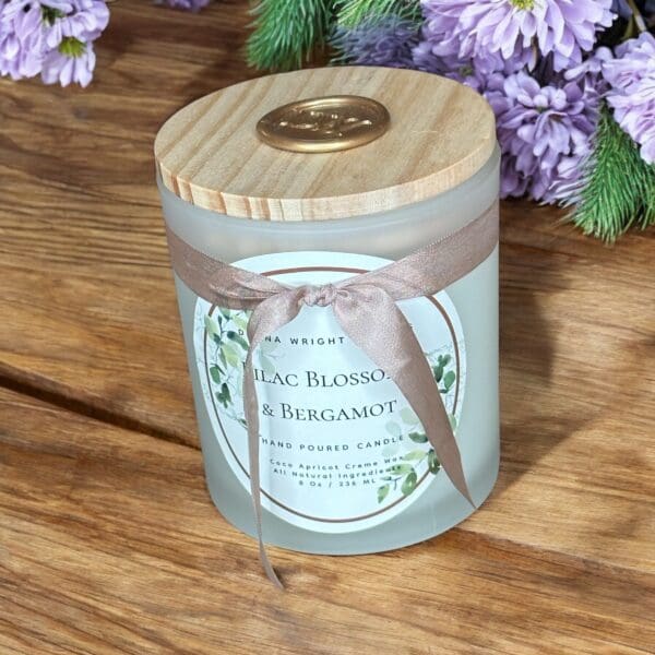 A frosted glass candle with a wooden lid and a ribbon bow sits on a wooden surface. The candle's label reads "Lilac Blossom & Bergamot." In the background, purple flowers and green foliage are visible.