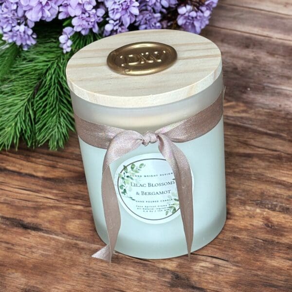 A frosted glass jar candle with a wooden lid and a bronze-colored ribbon is labeled "Lilac Blossoms & Bergamot." The candle rests on a wooden surface, surrounded by sprigs of green pine and a bouquet of purple flowers.