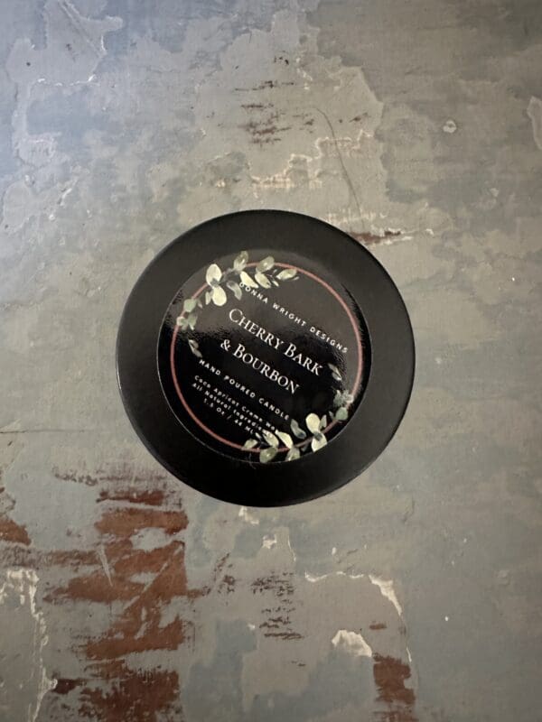 A round black candle labeled "Cherry Bark & Bourbon" by Stone Briar Designs is placed on a distressed, rustic surface with gray and brown tones. The 1.5 Oz candle label features a botanical design with leaves on a dark background, evoking the warm scents of spiced orange pomander.