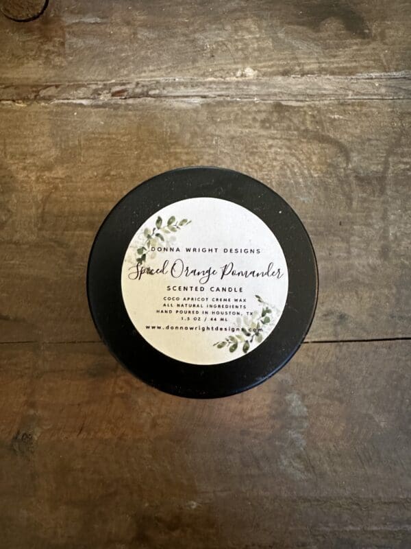 A black-lidded candle tin displayed on a wooden surface. The label reads "Donna Wright Designs – Spiced Orange Pomander Candles. Scented Candle. Coco Apricot Candle Wax. All natural ingredients. Hand poured in North Texas." The label has a small green leaf design and the tin holds 1.5 Oz of wax.