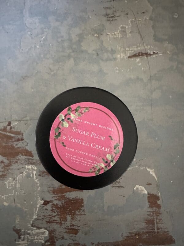 A hand-poured, 1.5 Oz candle with a black lid sits on a distressed, painted surface. The label reads "Sugar Plum & Vanilla Cream" and is adorned with green leaves and small red berries. The brand name, "Emma Wright Designs," is also visible on the label. Pair it with our Spiced Orange Pomander candles for a festive vibe.