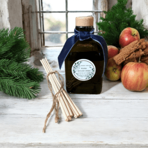 Warm Mulled Cider Diffuser by Donna Wright Designs on a white wooden table with pine, apples and cinnamon in the background with an open window behind.