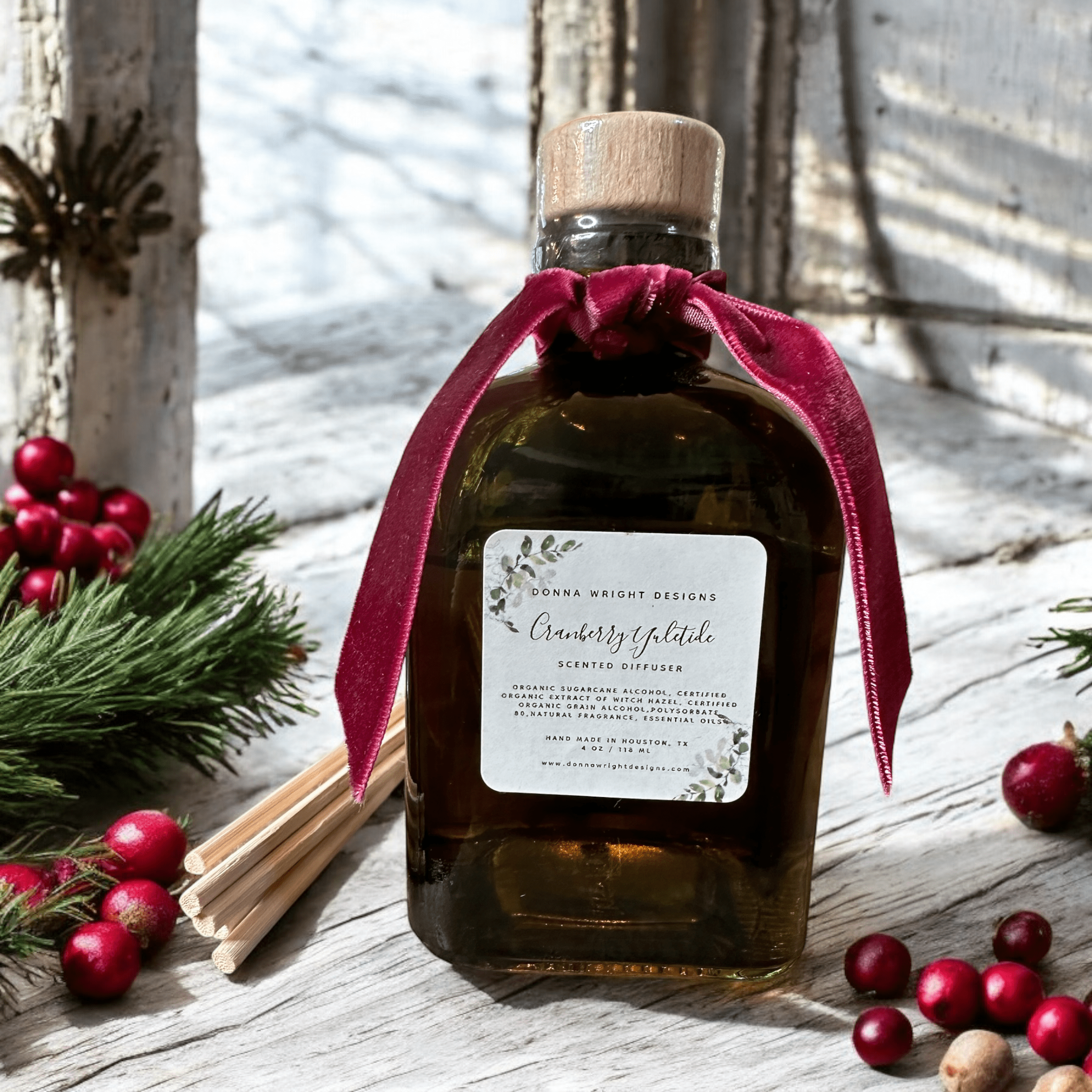 A glass bottle filled with scented diffuser liquid labeled "Cranberry Vybtlake" from Donna Wright Designs. The bottle is adorned with a burgundy ribbon and surrounded by cranberries, pine sprigs, and reed sticks on a rustic wooden surface.