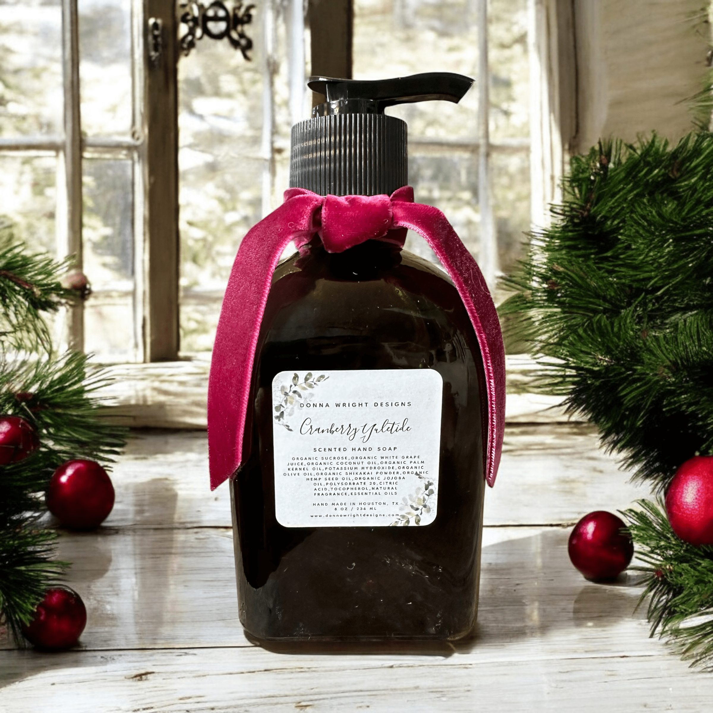 A dark brown bottle of scented hand soap with a pump lid is adorned with a pink velvet ribbon. It sits on a wooden windowsill surrounded by green pine garlands with red ornaments. Sunlight filters in through the window, illuminating the festive scene.