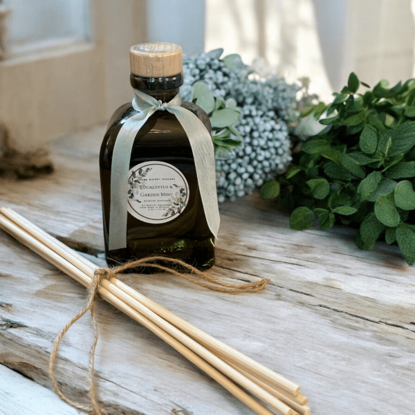 A decorative Eucalyptus & Garden Mint Reed Diffuser with a wooden cap rests on a wooden surface. Embellished with a ribbon, it is accompanied by several reed diffuser sticks tied with twine. Sprigs of eucalyptus and mint create a serene backdrop.