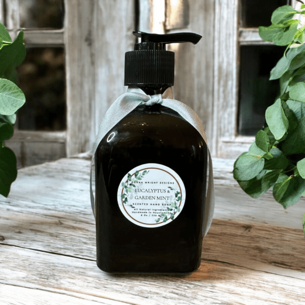 A dark green bottle of Eucalyptus & Garden Mint Hand Soap rests on a rustic wooden surface, surrounded by greenery and framed by a weathered window backdrop. The pump bottle is adorned with a light green ribbon tied around its neck.