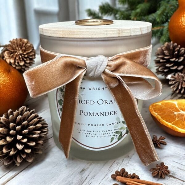 A frosted glass candle jar with a wooden lid is adorned with a cream and brown velvet ribbon tied in a bow. The label reads "Spiced Orange Pomander." Surrounding the candle are oranges, pinecones, and a slice of dried orange, creating a festive atmosphere.