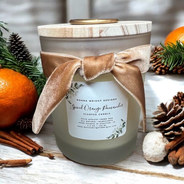 A glass jar candle labeled "Spiced Orange Pomander" is adorned with a beige velvet ribbon. The candle is surrounded by whole oranges, pinecones, cinnamon sticks, and greenery, all placed on a wooden surface, creating a rustic and festive atmosphere.