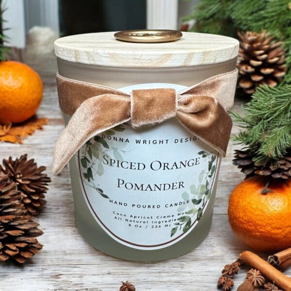 A hand-poured candle in a frosted glass jar labeled "Spiced Orange Pomander" by Donna Wright Design sits among oranges, pine cones, and evergreen branches. The candle features a wooden lid and a beige velvet bow around the jar.