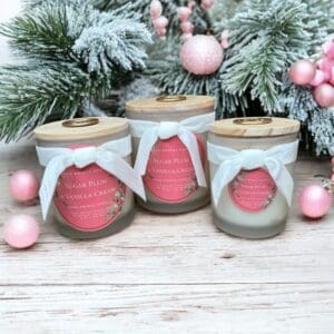 Three decorative candles with wooden lids and white bows are placed on a wooden surface. The candles have labels reading "Sugar Plum & Vanilla Cream." Pastel pink ornaments and snow-covered pine branches are arranged behind the candles, creating a festive holiday scene.