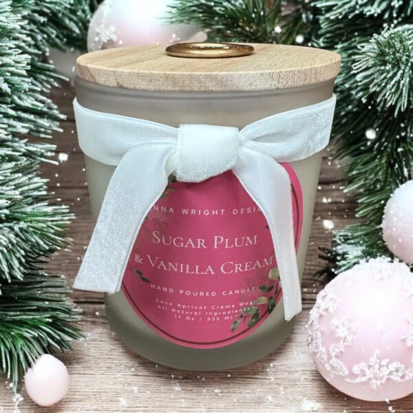 A frosted glass candle with a wooden lid is tied with a white ribbon. The label reads "SUGAR PLUM & VANILLA CREAM." Surrounding the candle are green pine branches and pink ornaments, evoking a festive, holiday theme.