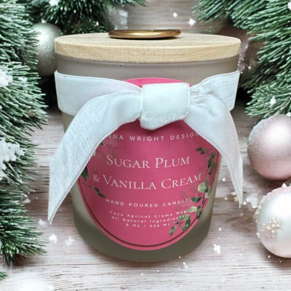 A scented candle in a frosted glass jar with the label "Sugar Plum & Vanilla Cream." The jar is tied with a white ribbon and surrounded by evergreen branches and Christmas ornaments. The product is branded as "Anna Wright Designs.