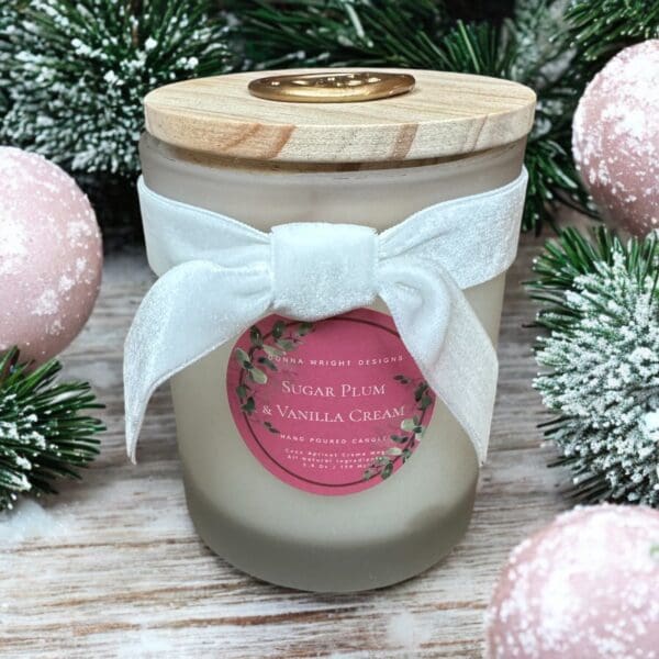 A frosted holiday candle with a wooden lid and a white velvet bow. The red label reads "Sugar Plum & Vanilla Cream." The candle is surrounded by snowy pine branches and pink ornaments, set on a wooden surface.