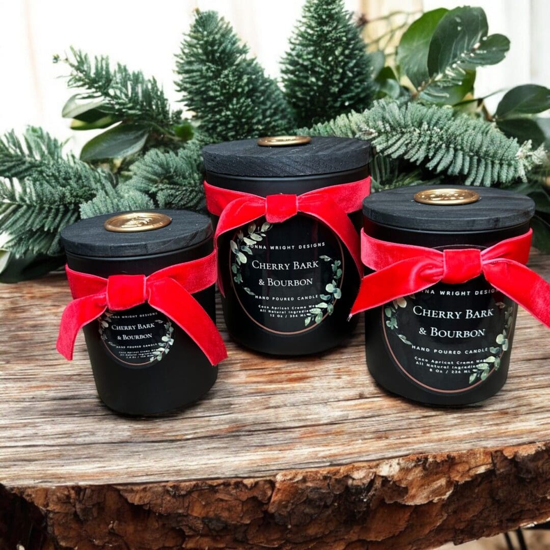 Three black candles labeled "Cherry Bark & Bourbon" are wrapped with red velvet ribbon and set on a rustic wooden surface adorned with evergreen branches. The candles vary in size, each with a gold seal on the matte black lid, creating a festive atmosphere reminiscent of the best fall and holiday markets.