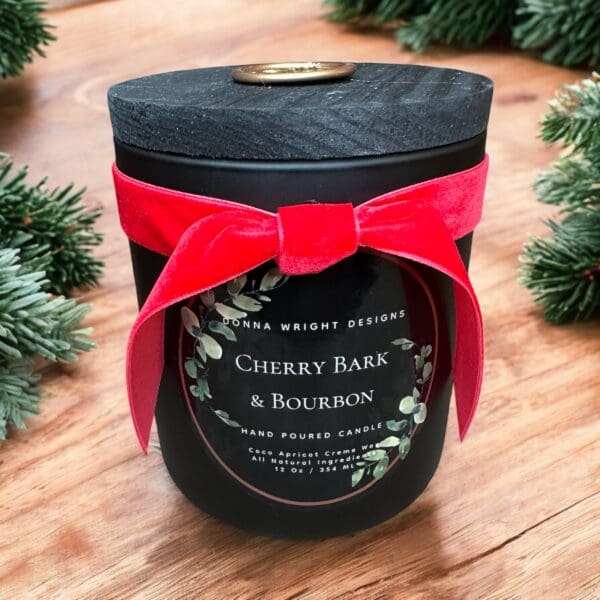 Black candle jar with a wooden lid and a red velvet bow, labeled "Cherry Bark & Bourbon" from Donna Wright Designs. The candle is surrounded by pine branches on a wooden surface. Text notes it's hand-poured with natural ingredients.