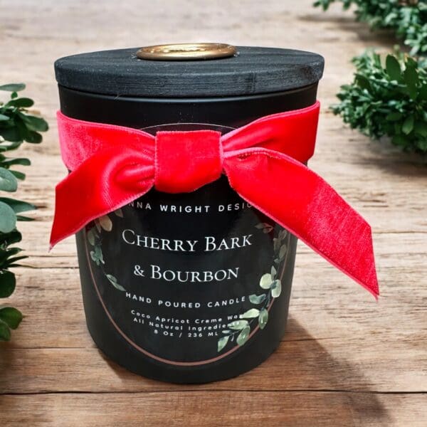 A black candle jar with a wooden lid, adorned with a vibrant red ribbon tied in a bow. The label reads "CHERRY BARK & BOURBON," indicating a hand-poured candle with coco apricot creme wax and all-natural ingredients. The jar sits on a wooden surface.