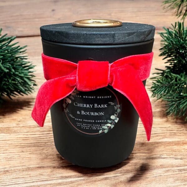 A black candle jar with a lid is adorned with a large red velvet bow. The label on the jar reads, "Cherry Bark & Bourbon, Hand Poured Candle." The candle is placed on a wooden surface with small green pine branches on the sides.