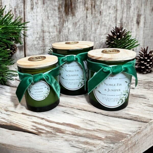 Three green glass candles of varying sizes sit on a rustic wooden surface. Each candle has a light wooden lid and a green ribbon tied into a bow around the neck. Nearby are pine branches and pinecones, enhancing the festive, natural atmosphere.