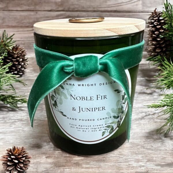 A green candle in a glass jar with a wooden lid is adorned with a green velvet ribbon. The label reads "Noble Fir & Juniper" and "Hand Poured Candle" among greenery decorations. Pinecones and pine branches surround the candle on a wooden surface.