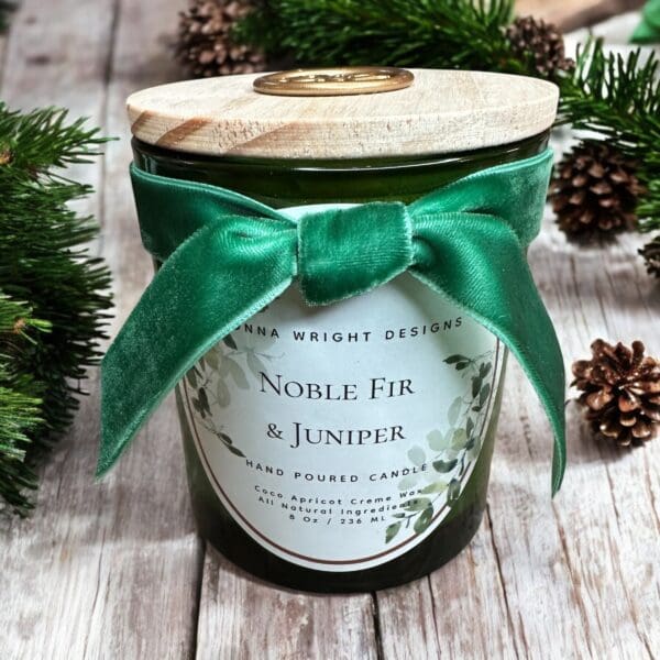 A candle with a wooden lid, decorated with a green velvet bow, labeled "Noble Fir & Juniper" by Anna Wright Designs. The jar is surrounded by pinecones and green pine branches, sitting on a wooden surface, creating a festive holiday ambiance.