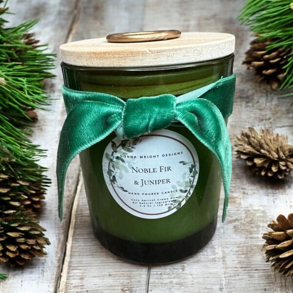 A green candle in a glass jar with a wooden lid is adorned with a green velvet bow. The label reads "Noble Fir & Juniper" by "Donna Wright Designs." The candle is surrounded by pinecones and fir branches on a rustic wooden surface.