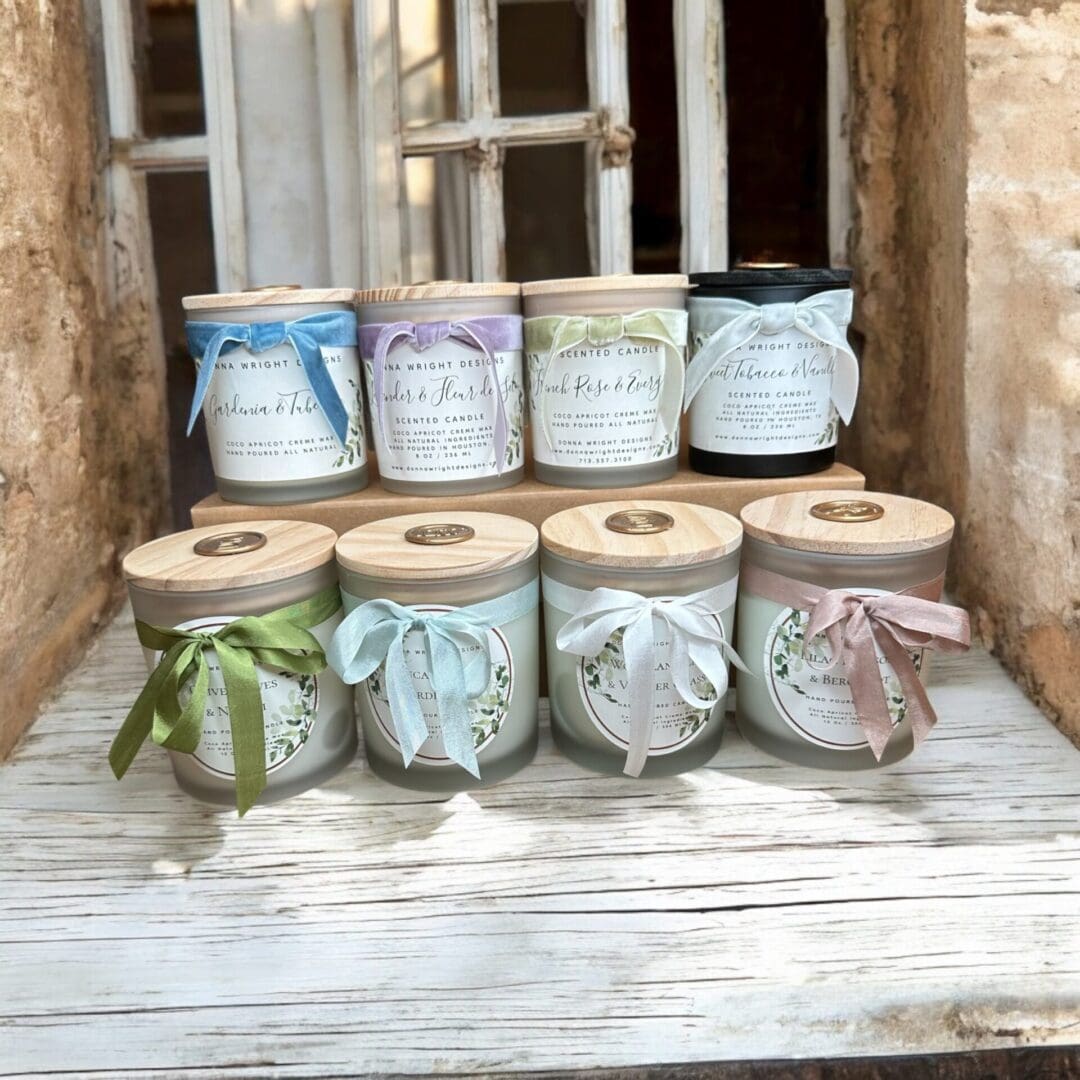 A Donna Wright Designs signature collection of nine scented candles is arranged on a wooden surface. Each candle, in glass jars with various colors, is topped with a wooden lid and adorned with a colorful ribbon. They are placed in front of a rustic window with peeling paint.