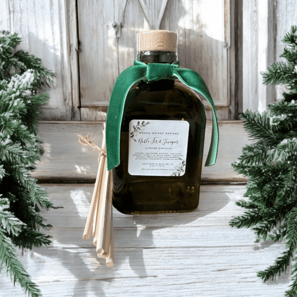 A Christmas-themed reed diffuser with a clear bottle, green liquid inside, and a green ribbon around the neck. It is placed against a white wooden background with green pine branches framing the image on the left and right sides.