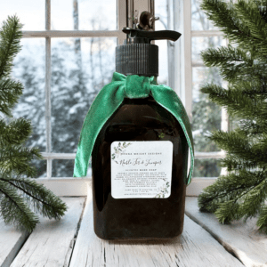 A bottle of hand soap with a green ribbon tied around its neck stands on a white wooden surface. The label reads "Mistle-Thiough Joy-Seize," and displays a festive design. The background shows a snowy outdoor scene through a window, with evergreen branches on the sides.