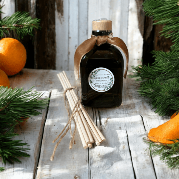 The Spiced Orange Pomander Reed Diffuser sits on a rustic wooden surface surrounded by evergreen branches and spiced oranges. The bottle has a ribbon tied around its neck and is accompanied by several reed sticks. The background features a weathered wooden wall, evoking the warmth of a handmade pomander.