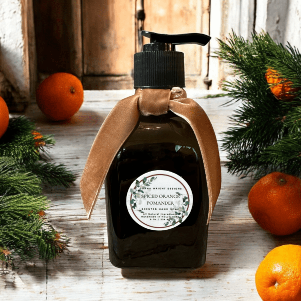 A bottle of Spiced Orange Pomander Hand Soap with a pump dispenser and adorned with a brown ribbon bow is set on a wooden surface. The background features evergreen branches, small oranges, and a rustic wooden door, creating a festive atmosphere.