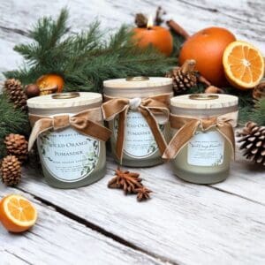 Three scented candles labeled "Spiced Orange Pomander" with beige ribbons are on a white wooden surface. They are surrounded by pine branches, pine cones, whole and sliced oranges, and a lit candle in the background, creating a festive, cozy atmosphere.