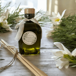 A green glass bottle with a wooden cork and a white ribbon around its neck is placed on a wooden surface. The label reads "Eucalyptus & Garden Mint Reed Diffuser." Surrounding the bottle are white lilies, some reed diffuser sticks, and sprigs of eucalyptus.