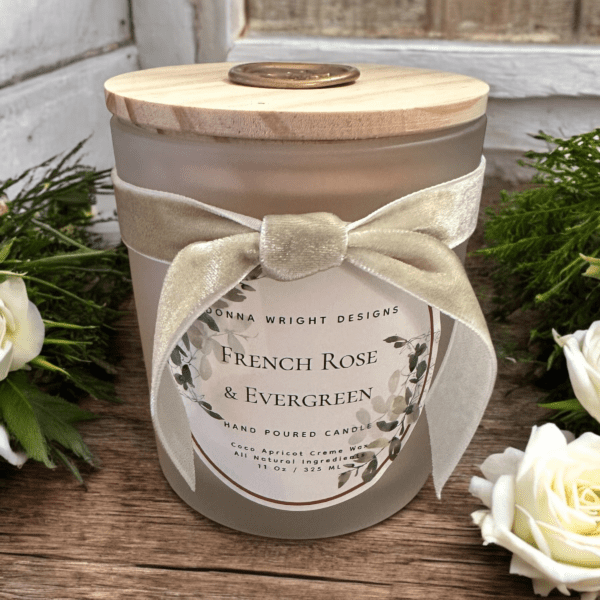 A glass jar candle from Donna Wright Designs labeled "French Rose & Evergreen" features a wooden lid and a cream ribbon, nestled amidst lush greenery and white roses on a rustic wooden surface.