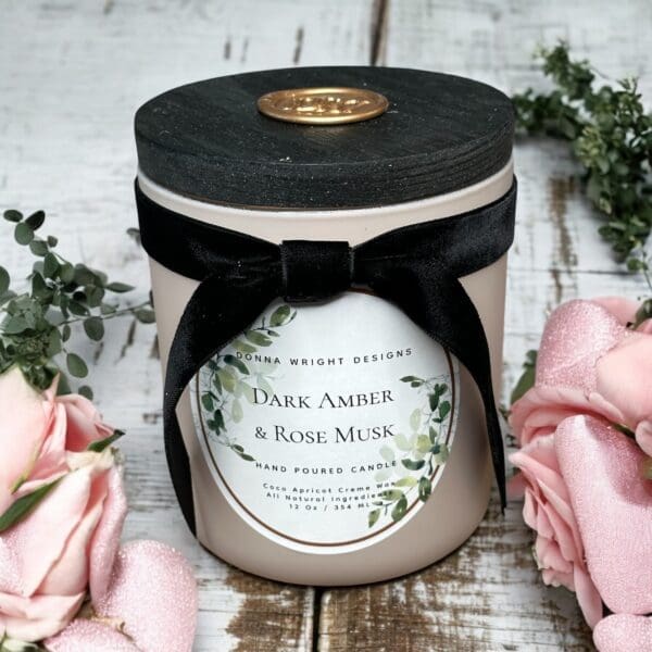 A "Dark Amber & Rose Musk Candles" scented candle with a wooden lid and black ribbon rests on rustic wood. The air is rich with rose musk aroma, enhanced by the elegance of surrounding pink roses.