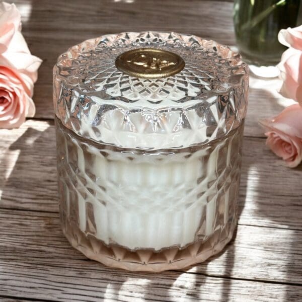 The Dark Amber & Rose Musk Candle, with a textured glass design and gold-embellished lid, sits on a wooden surface exuding a rose musk aroma. Nearby, pink roses are softly illuminated by natural light.