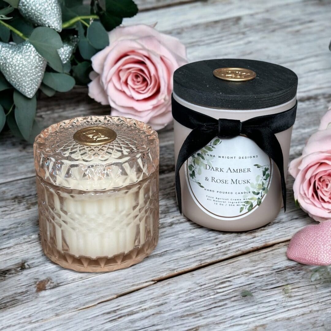 A decorative candle in a faceted glass jar accompanies a "Dark Amber & Rose Musk" lidded candle. Their delicate scent enhances the display of pink roses and eucalyptus on the rustic wooden surface.