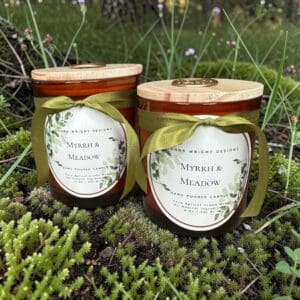 Two amber jars labeled “Myrrh & Meadow Candles,” each adorned with a green ribbon and topped with a wooden lid, rest on mossy forest ground among grass and delicate purple blooms—an inviting scene that perfectly reflects the natural tranquility these Luxury Myrrh & Meadow Candles embody.