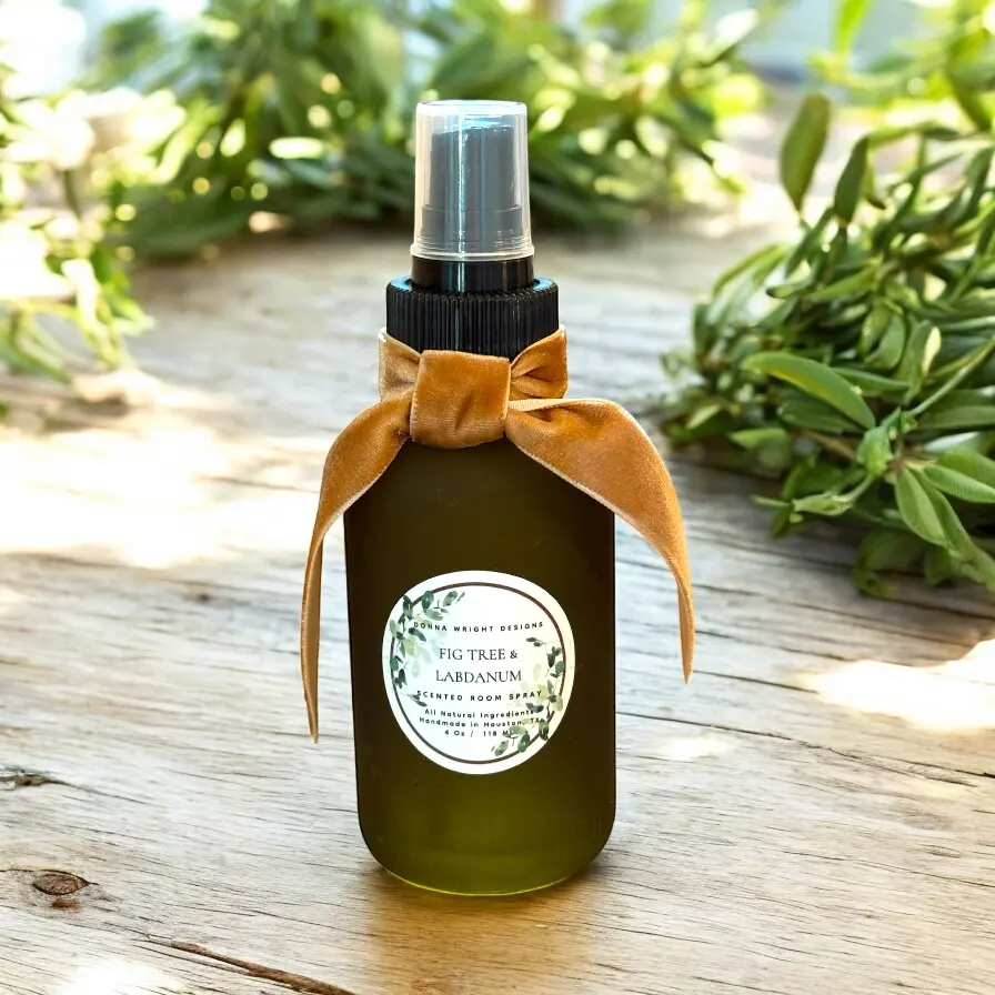 A green glass Fig Tree & Labdanum Room Spray bottle with a gold velvet ribbon and round label sits on a wooden surface, framed by green foliage in the background.