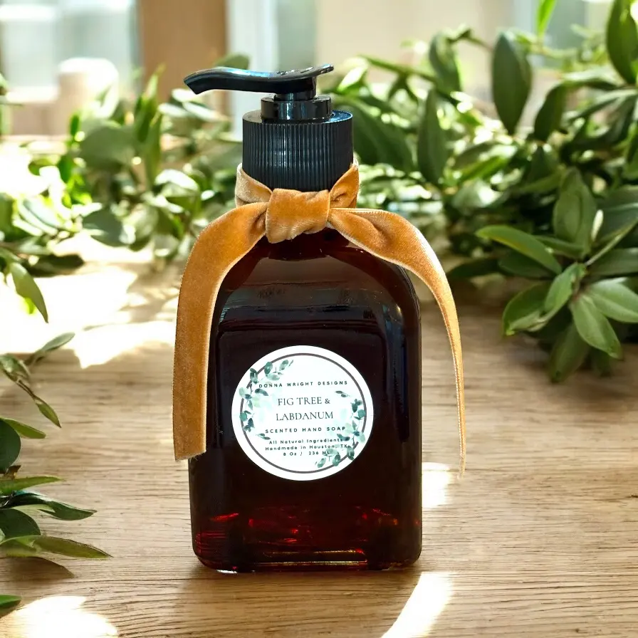 A dark amber soap dispenser wrapped in a gold velvet ribbon and labeled “Fig Tree & Labdanum” sits among green leaves in sunlight—pair with the Fig Tree & Labdanum Room Spray for a perfectly matched fragrance duo.