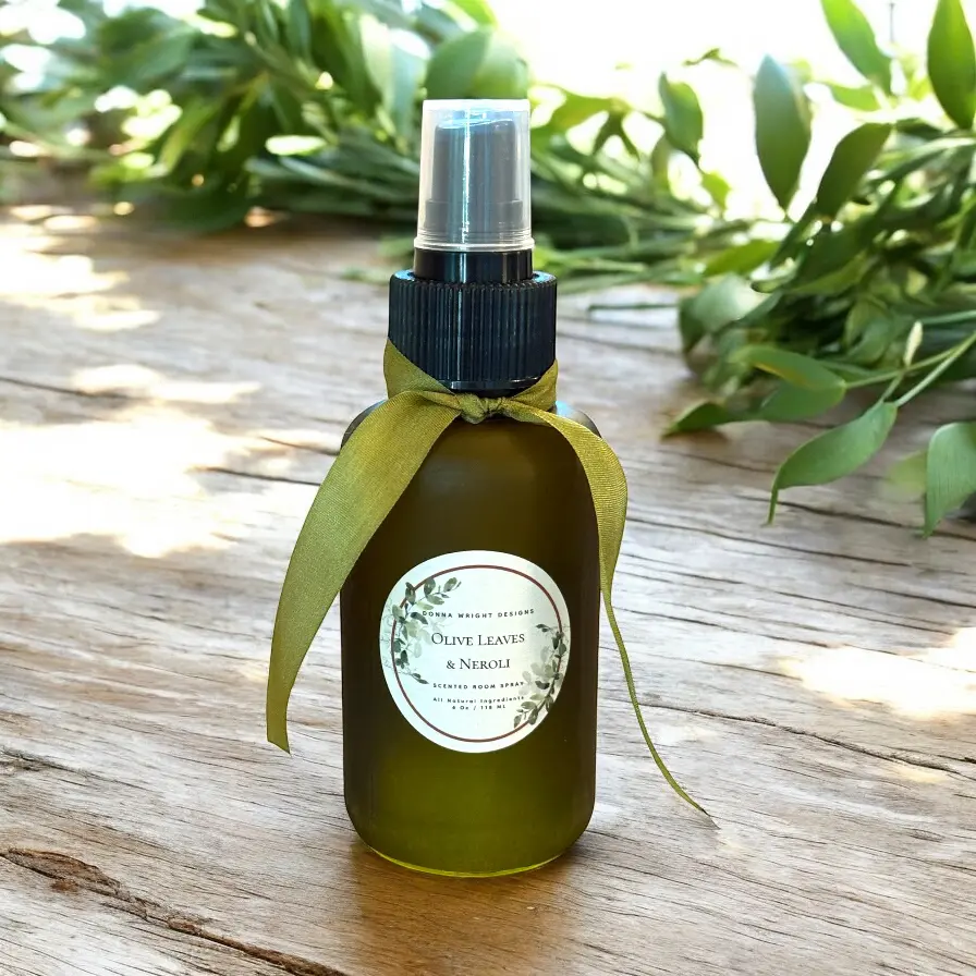 A Olive Leaves & Neroli Room Spray sits on a wooden surface adorned with a green ribbon, set against blurred leafy greenery in the background.