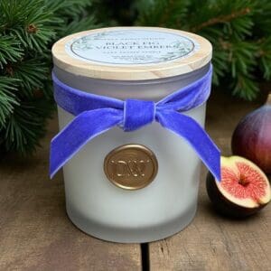 A frosted glass Black Fig & Violet Embers Candle with a wooden lid, blue velvet ribbon, and wax seal sits on wood near figs and pine branches. The label displays “Black Fig & Violet Embers Candles.”.
